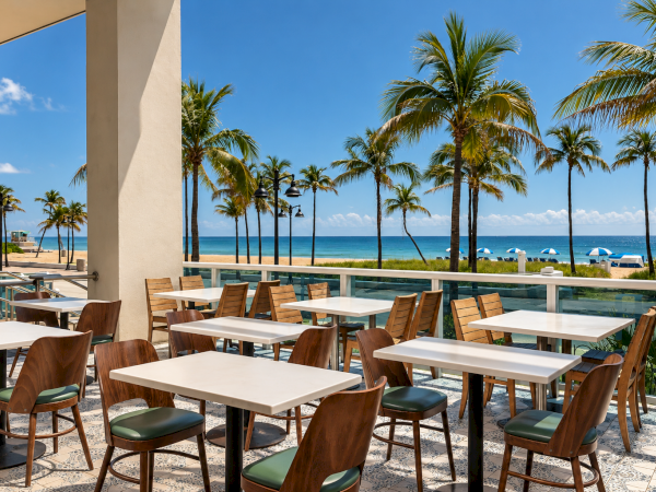Sunlit seaside patio with wooden tables, green chairs, palm trees, blue ocean, and umbrellas under a clear sky.
