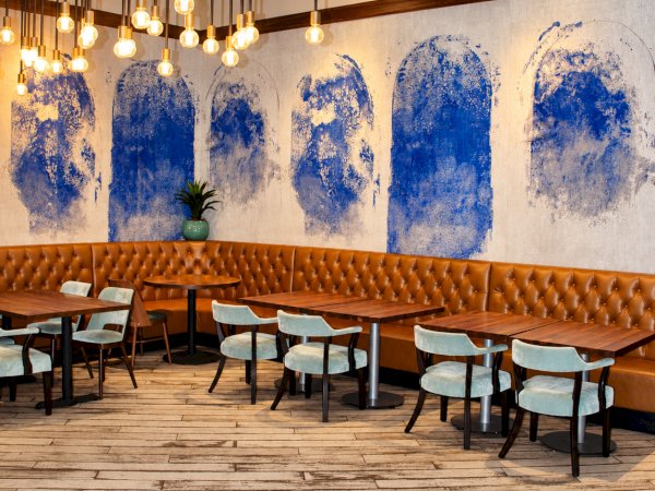 Cozy restaurant interior with tufted leather banquette, wooden tables, hanging warm bulbs, and blue abstract wall art.