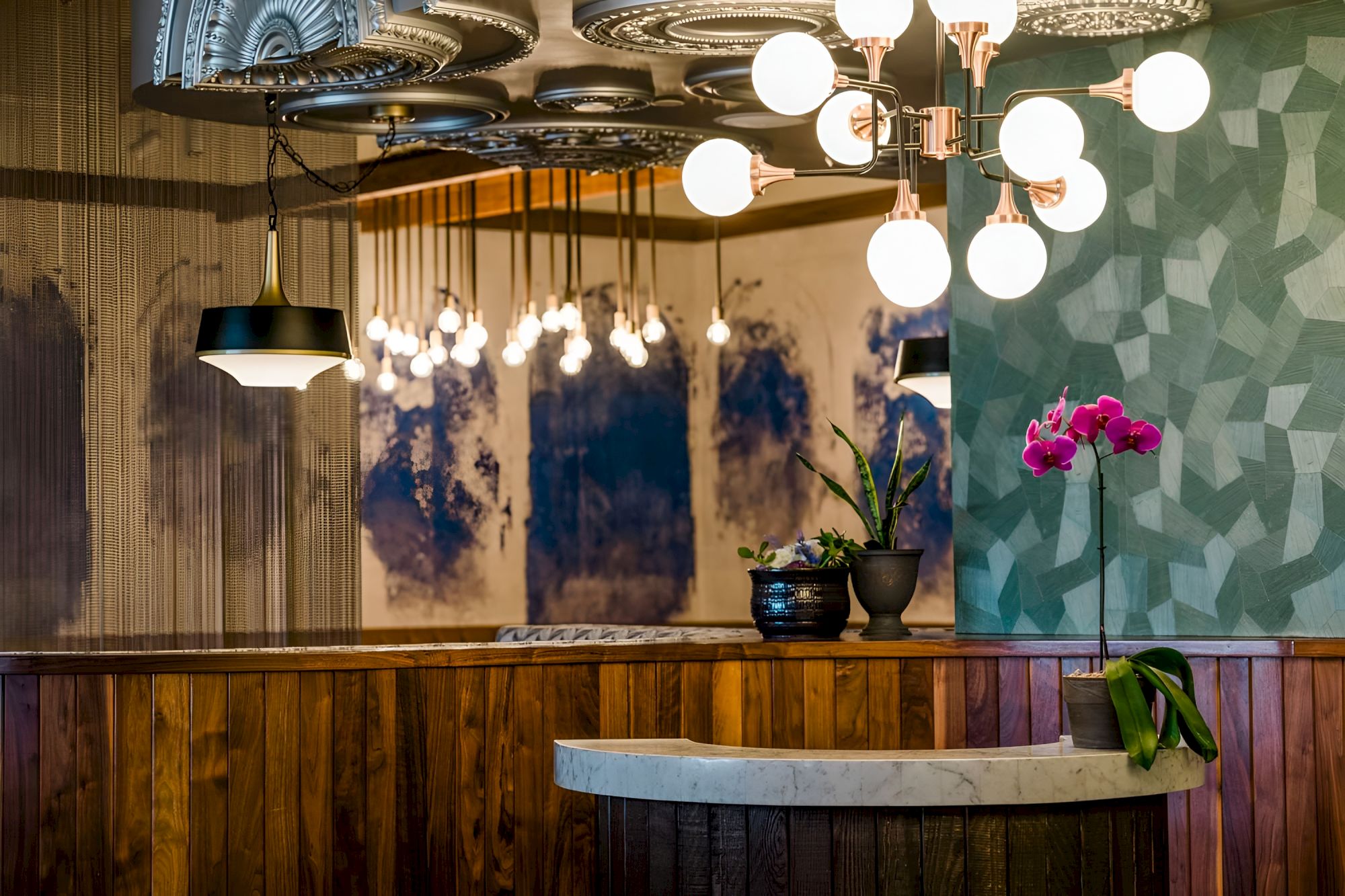 A stylish lounge interior with ornate ceiling lights, gold accents, patterned walls, and a small table vase of pink orchids.