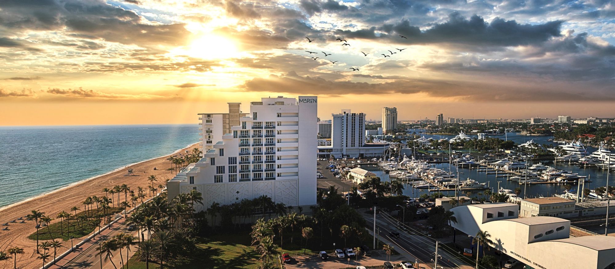 A seaside resort overlooks a marina with many yachts at sunset, while birds fly through dramatic clouds.