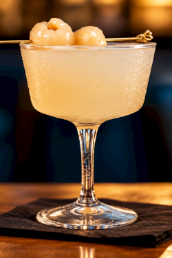 A golden cocktail in a coupe glass with two creamy spheres on top and a lemon twist, garnished with a skewer.