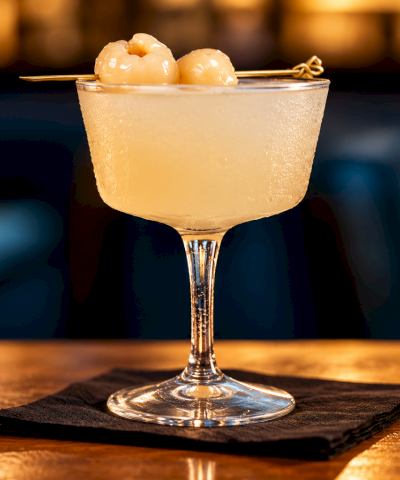 A golden cocktail in a coupe glass with two creamy spheres on top and a lemon twist, garnished with a skewer.