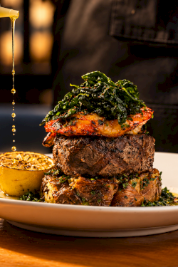 A chef pours golden oil over a herb-topped steak on a plate, beside a halved lemon, in a sizzling kitchen scene.