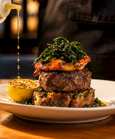 A chef pours golden oil over a herb-topped steak on a plate, beside a halved lemon, in a sizzling kitchen scene.