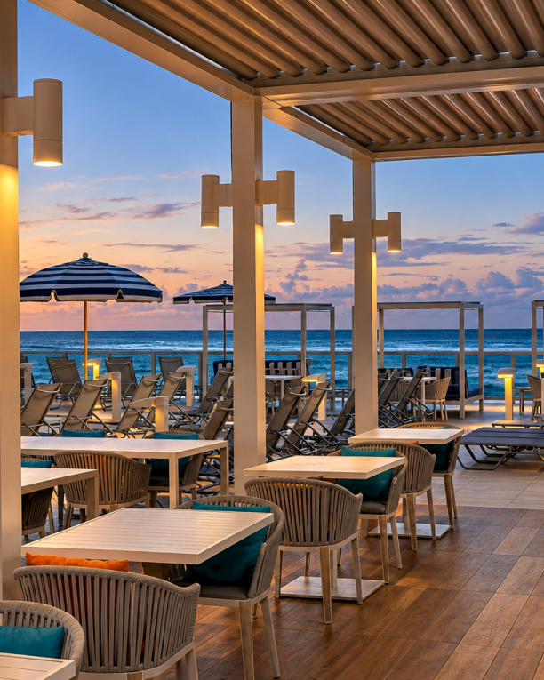A luxury oceanfront terrace with wooden loungers, umbrellas, and warm lights at sunset over the blue sea.