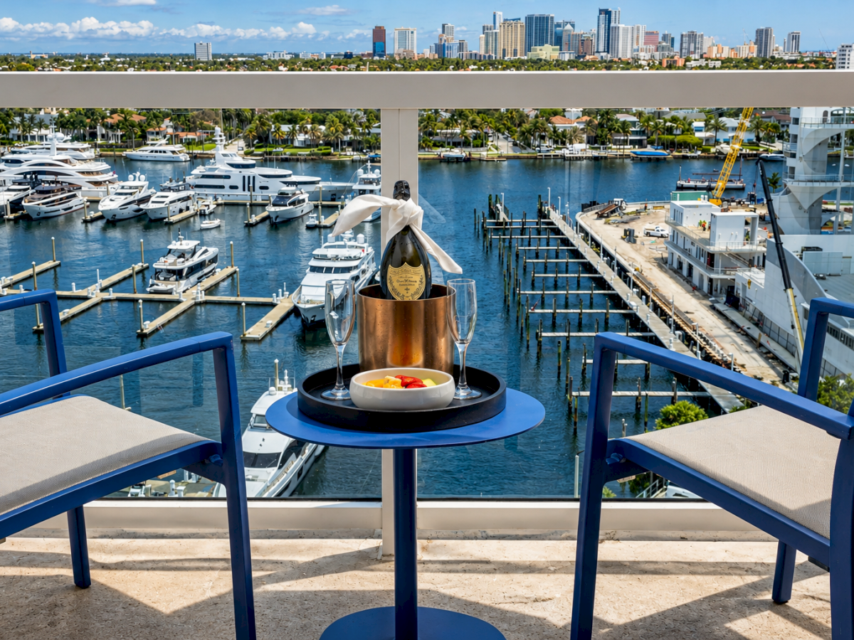 Yachts in a marina with a champagne bucket and glasses on a table beside blue chairs, facing a city skyline under clouds.