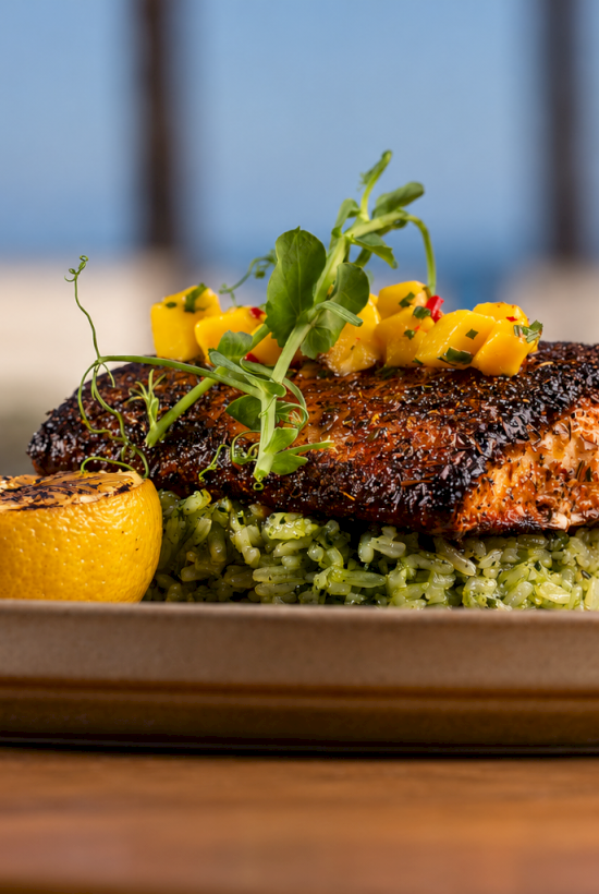 A char-grilled salmon steak on rice with chopped yellow cheese, microgreens, and a lemon wedge on the side.