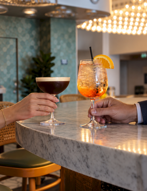 Two people clink cocktail glasses&mdash;one dark and one orange citrus iced drink&mdash;in a cozy, chandelier-lit lounge, smiling in the moment!.