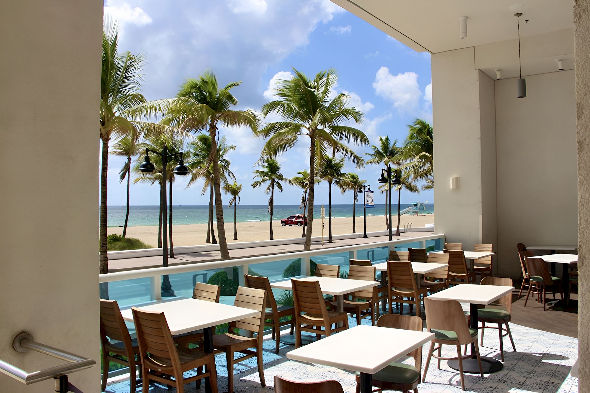 Sunlit patio with tables and chairs overlooking palm trees, ocean, and a sandy beach under blue sky.