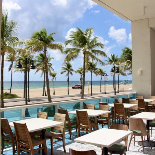 A beachfront patio restaurant with white tables, chairs, palm trees, and a view of the ocean under a bright sky.