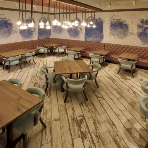 A cozy caf&eacute; lounge with wooden tables, green chairs, tufted brown booths, blue mural walls, and bright hanging pendant lights.