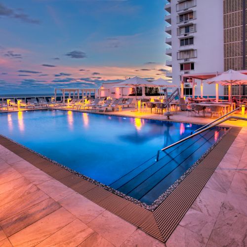 A rooftop hotel pool at sunset with deck chairs and umbrellas, beside tall apartment-style buildings under a colorful sky.