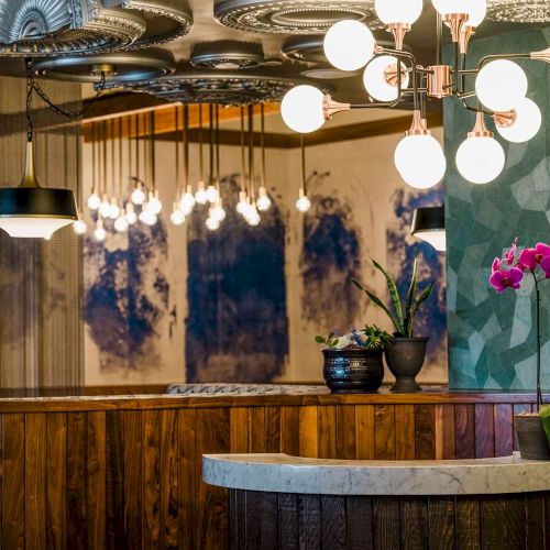 A stylish lounge interior with an ornate chandelier, warm lighting, a wooden bar counter, and bright pink orchids in a vase.