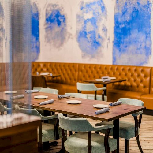 A modern restaurant dining area with wooden tables, upholstered green chairs, pendant lights, and blue-white wall art panels.