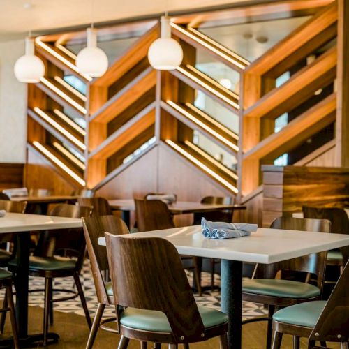 A modern restaurant interior with set tables and chairs beneath a glowing wooden staircase and hanging white pendant lights, beautifully lit.