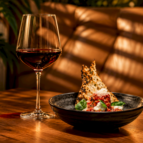 A glass of red wine sits beside a bowl of pizza-topped food on a table with warm candlelight.