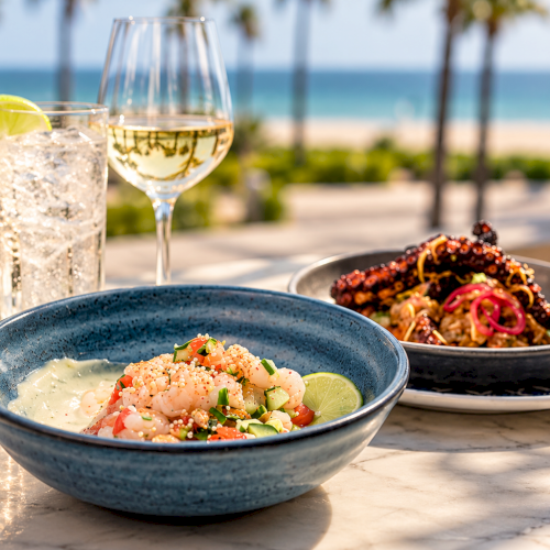 A sunny beachside table with shrimp ceviche and tacos, plus lime water and a cocktail glass, by the ocean.