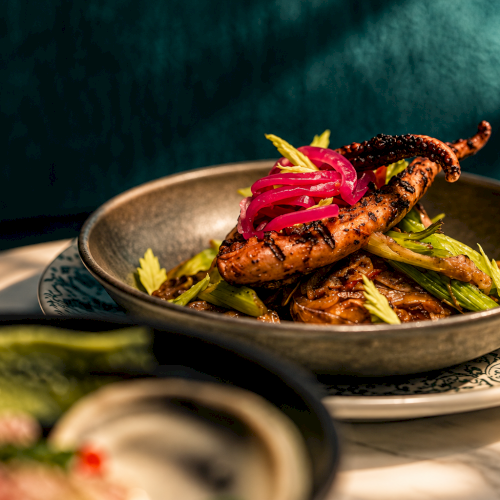 Grilled sausage on a plate with saut&eacute;ed greens and pickled red onions, garnished with sauce and herbs.