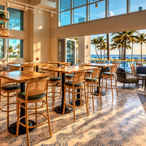 A bright tropical restaurant with wooden tables and wicker chairs, opening to palm trees and the ocean beyond.