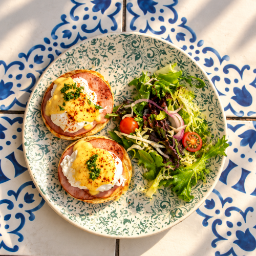 A plate of mixed greens salad with cherry tomatoes and two sunny-side eggs, on a blue-and-white tiled background.