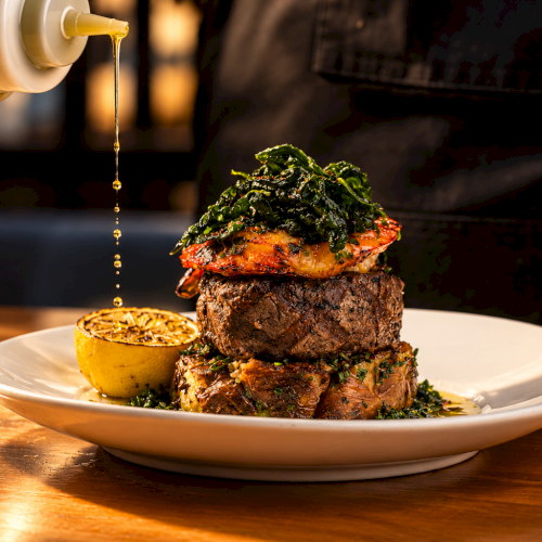 A chef pours golden sauce onto a grilled steak topped with greens on a plate, with a lemon half beside it!