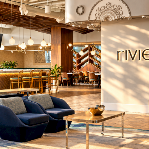 A stylish hotel lounge and bar interior with a &ldquo;rivera&rdquo; sign, seating, bar counter, pendant lights, and decorative wall arch.