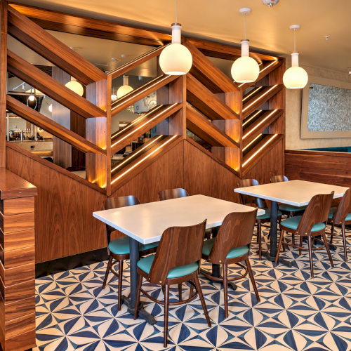 A modern restaurant interior with wooden wall partitions, warm pendant lights, tables, teal chairs, and patterned tile flooring.