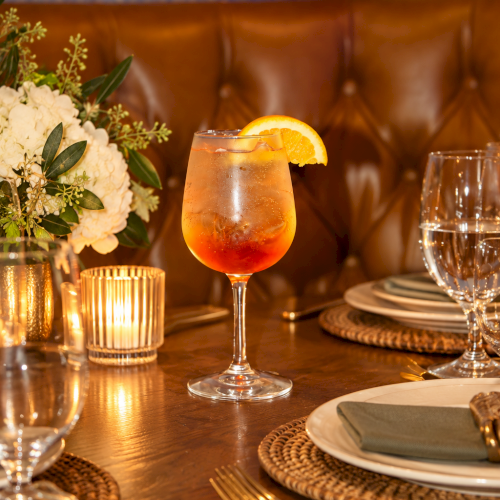 A sparkling orange cocktail in a stemmed glass sits on a styled table with flowers, cutlery, and wine glasses beside it.