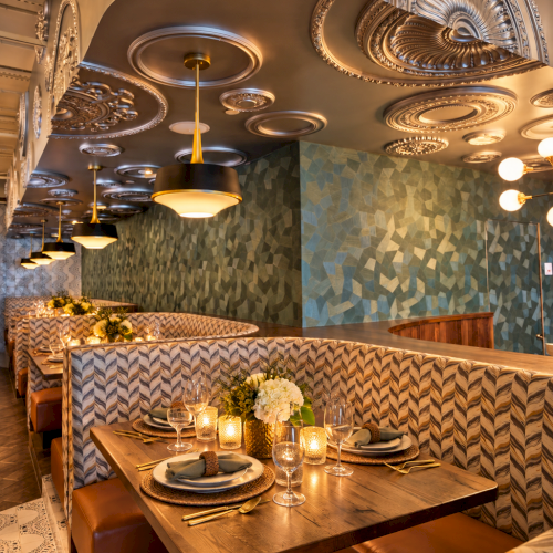 A stylish dining nook with ornate ceiling chandeliers, patterned banquette seating, and a set table glowing with warm light.