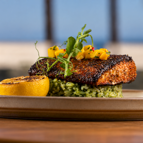 Plated grilled beef topped with herb sprouts and cheese, served over green rice, with a side lemon on a wooden plate.