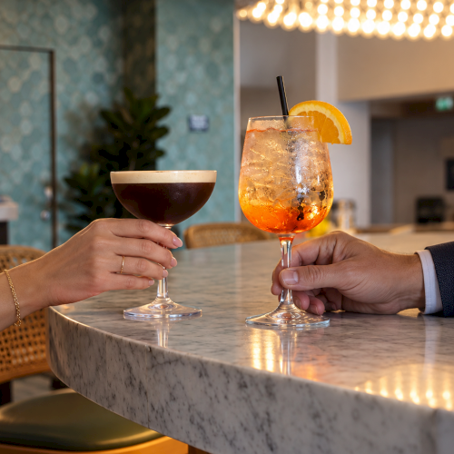 Two people clink glasses over a marble bar: a dark cocktail and an orange iced drink with straw.