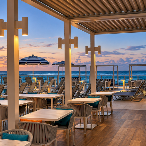 Oceanfront terrace at sunset with lounge chairs, striped umbrellas, and a pool, framed by a modern pergola and ocean waves.