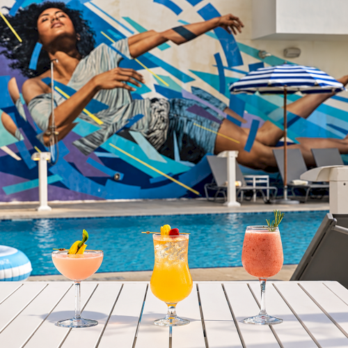 A sunny pool deck with three fruity cocktails in front, while a colorful mural backdrop shows a woman lounging on a daybed.